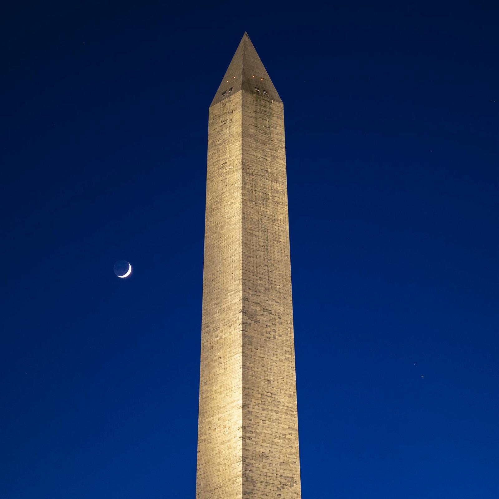 Floodlit obelisk against a deep blue sky with a crescent Moon and two close planets.