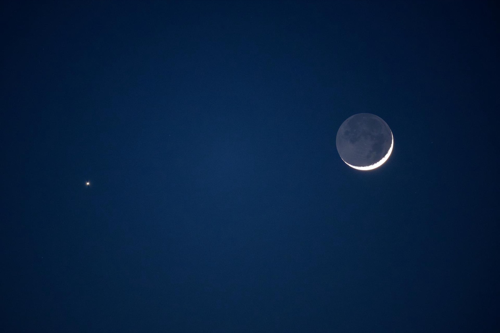 Crescent Moon with faint earthshine and Venus as a bright point to its left in a blue sky.