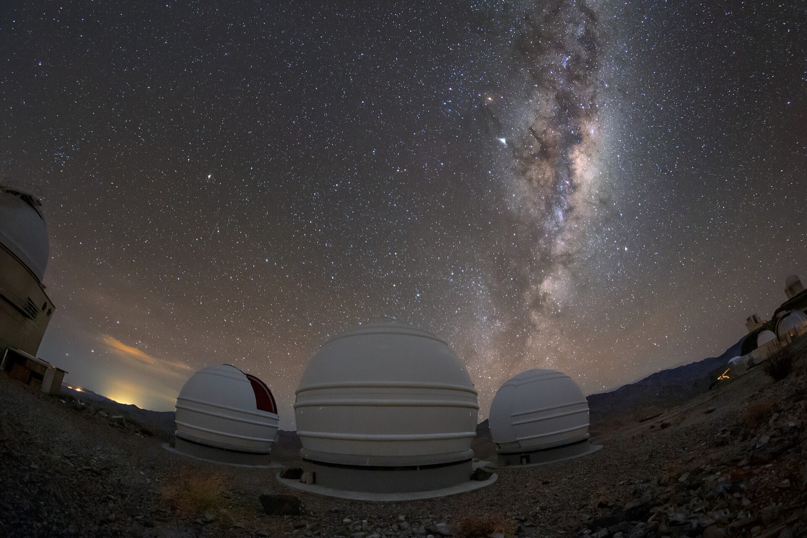 Milky Way rising above three white telescope domes on a desert ridge at night.