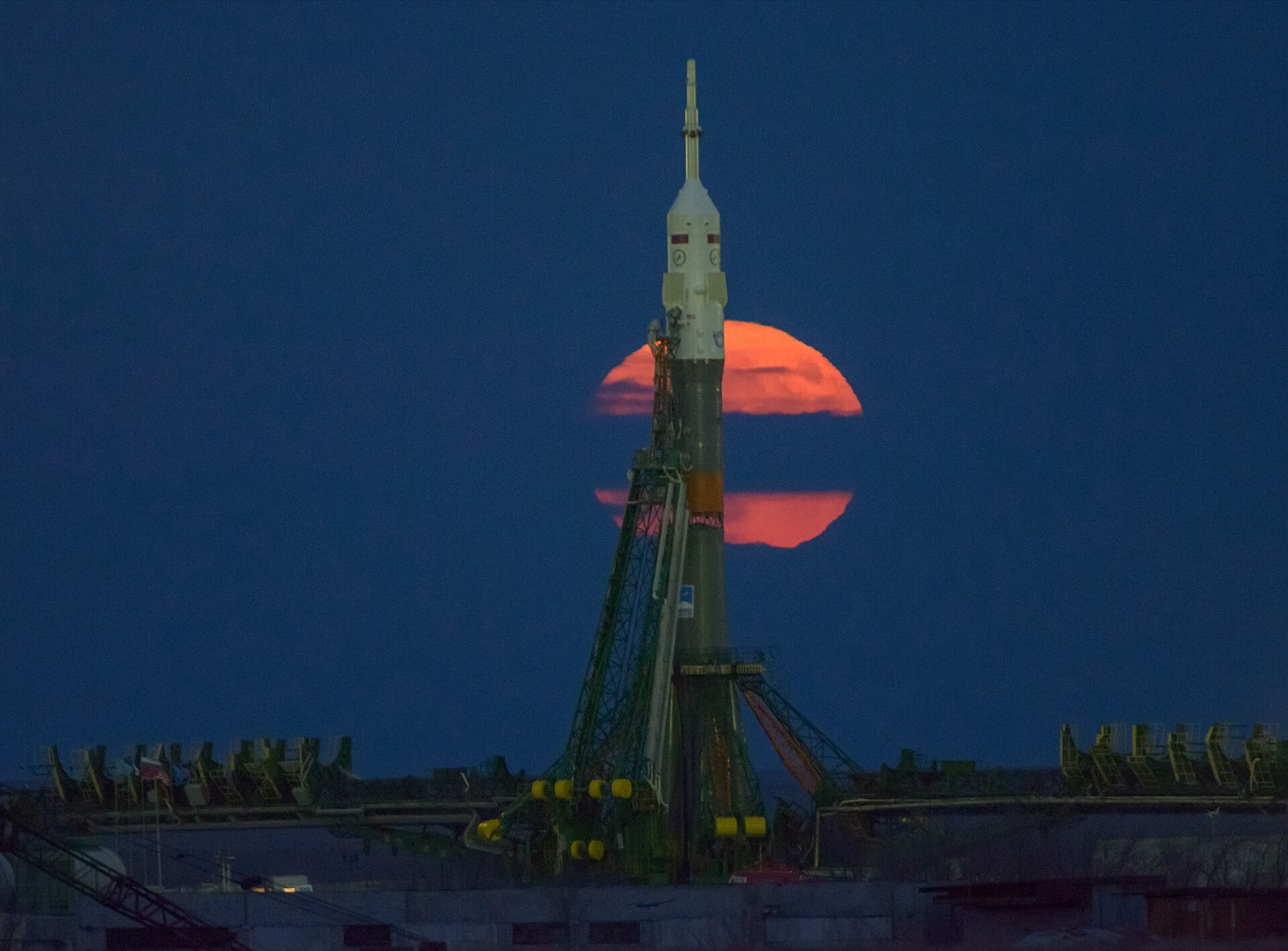 Orange full Moon rising behind a Soyuz rocket and launch tower at dusk.