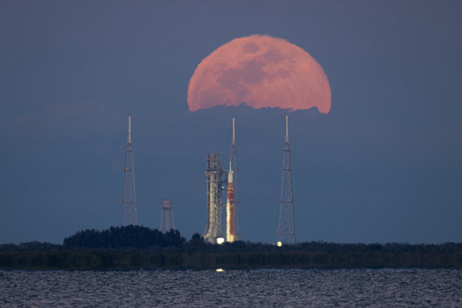Large pink full Moon rising behind a rocket and three lightning towers on a launch pad across water.