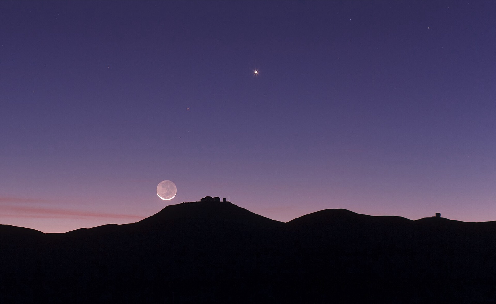 Thin crescent Moon with earthshine over a dark ridge in twilight, two planets above.