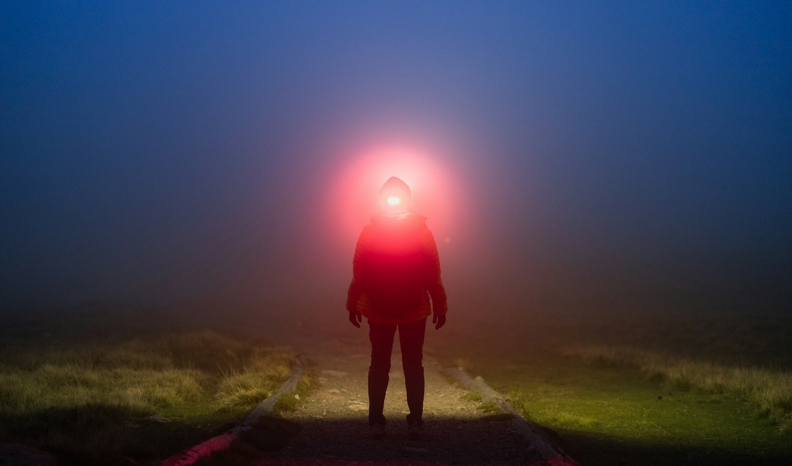 Silhouette of a person with a red headlamp glowing in fog on a dark mountain path.