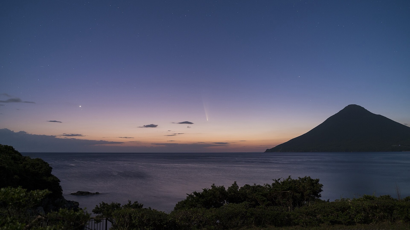 Faint comet with an upright tail in an orange and blue twilight sky beside a conical mountain over the sea.
