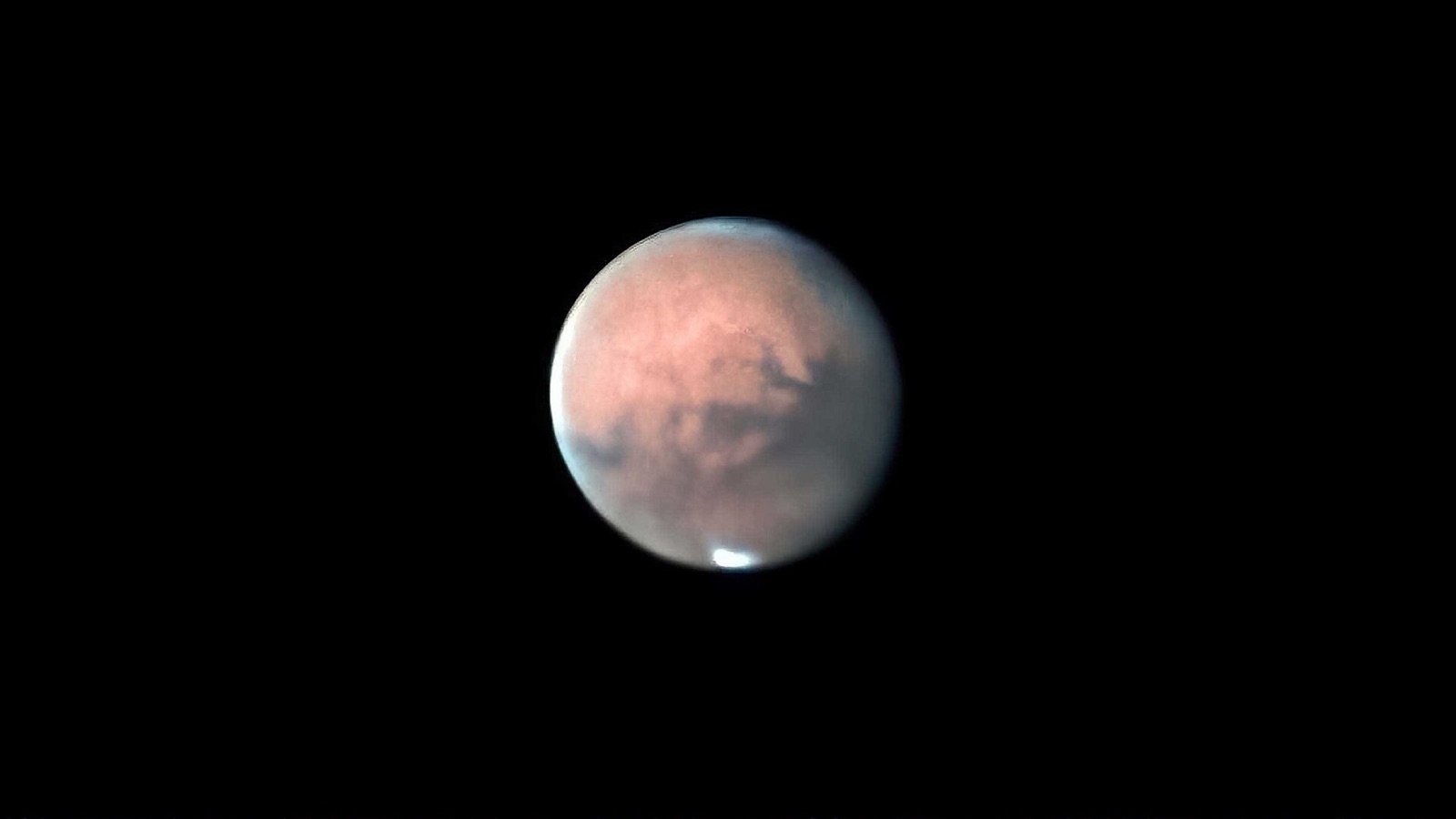 Orange disc of Mars with dark markings and a small white polar cap on a black background.