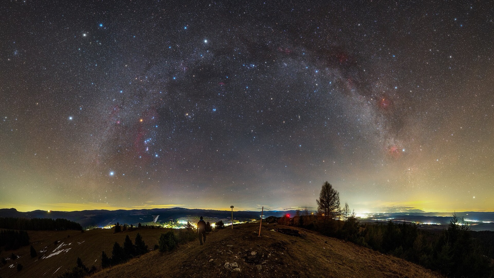 Wide panorama of the winter Milky Way and Orion above a lit valley landscape