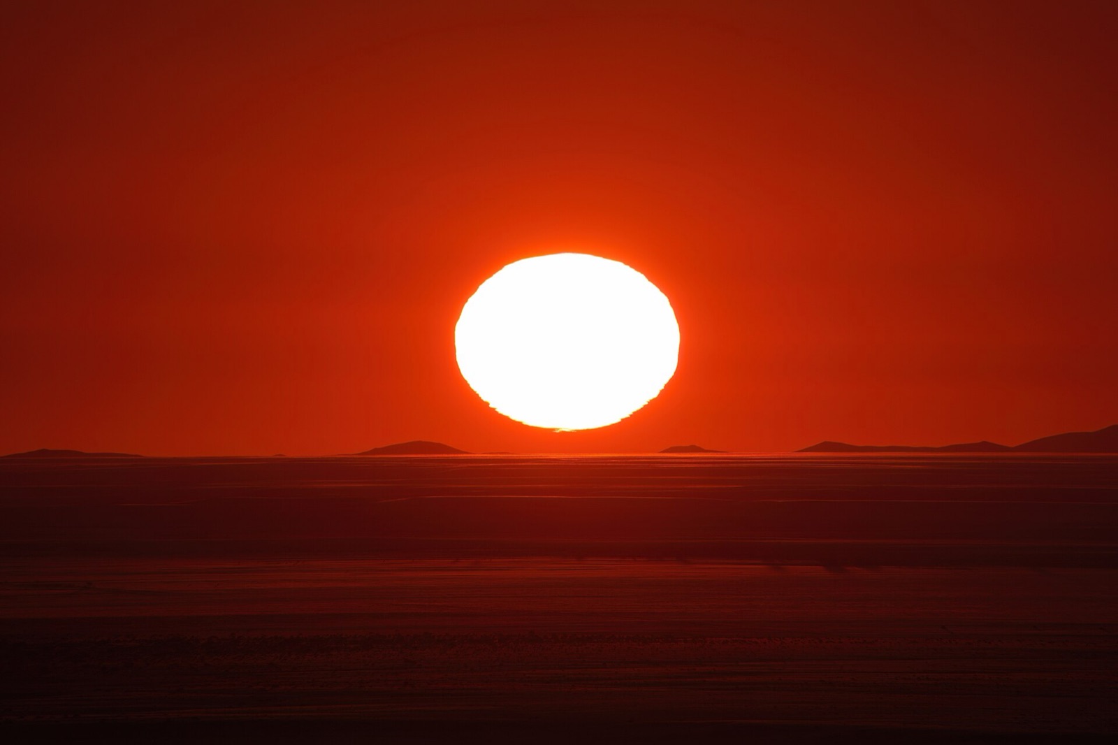 Flattened oval setting Sun with a wavy lower edge on a dark desert horizon under a red sky.