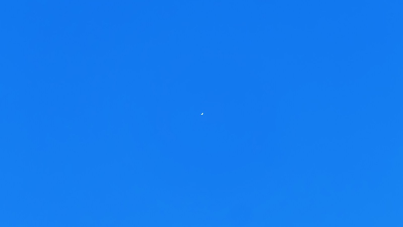 Plain blue daytime sky with Venus visible as a very small white crescent.